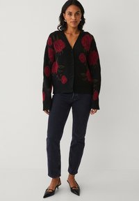 Zwarte cardigan met rode bloemenpatronen, v-hals en knoopsluiting. Gecombineerd met donkere jeans en zwarte puntige hakken.