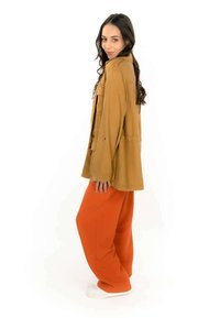 Veste surdimensionnée beige avec de longues manches, poches avant et coupe décontractée, accompagnée d'un pantalon large orange vif et de chaussures blanches.