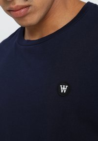 Námořnická modrá bavlněná tričko s kulatým výstřihem, krátkými rukávy a kruhovou černou nášivkou s bílým logem 'W' na levé straně hrudi.