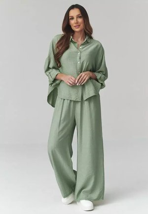Kobieta stojąca w luźnej zielono-szałwiowej koszuli na guziki i szerokich spodniach z białymi platformowymi butami na jasnym tle.