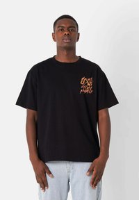 Camiseta de algodón negra con mangas cortas y un diseño de llamas coloridas en la parte izquierda del pecho. Lleva puestas jeans de mezclilla azul claro.