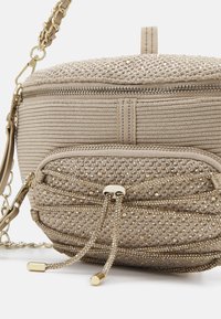 Béžová texturovaná kabelka přes rameno se zlatými zipy, krystalovými akcenty a řetízkovým popruhem. Obsahuje dvě přihrádky a dekorativní tkaničky.