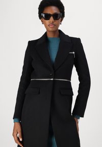 Cappotto nero su misura con chiusura a bottone, dettagli con zip argentata in vita e due tasche frontali. Sotto, una maglietta a coste di colore teal.
