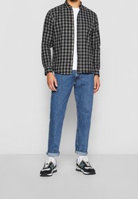 Camicia di flanella a quadri neri e grigi, t-shirt bianca sotto, jeans blu e sneakers nere con dettagli bianchi. Polsini dei jeans arrotolati.