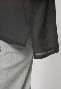 Tissu noir transparent avec détail d'ourlet fendu en angle sur un pantalon gris clair à fines rayures, montrant la texture du tissu et les coutures en gros plan.