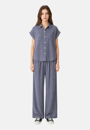 Femme debout face à l’avant, portant une chemise grise ample à manches courtes boutonnée, un pantalon large et des baskets noires et blanches sur fond blanc.