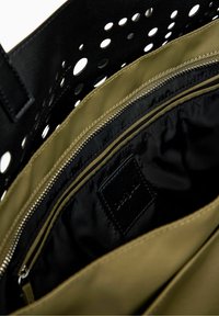 Sac cabas en cuir noir avec un motif ajouré en forme de points. L'intérieur est de couleur vert olive, avec un compartiment zippé noir et une petite étiquette logo.