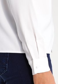 Primo piano di una persona che indossa una camicia bianca a maniche lunghe con polsini abbottonati e jeans di denim blu scuro.
