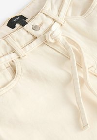 Pantalon en denim couleur crème avec un bouton, des passants pour ceinture, une poche avant et une ceinture en tissu nouée sous la taille avec une étiquette "NEXT".