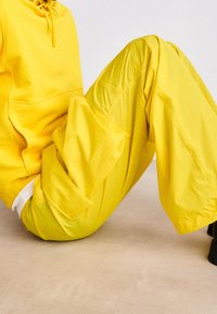 Geel hoodie en wijde broek gemaakt van zacht, glad materiaal, met elastische accenten en zijzakken, tegen een neutrale achtergrond.