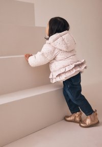 Veste rose pâle matelassée avec des pois dorés et un ourlet volanté, associée à un jean bleu foncé et des bottines dorées brillantes.