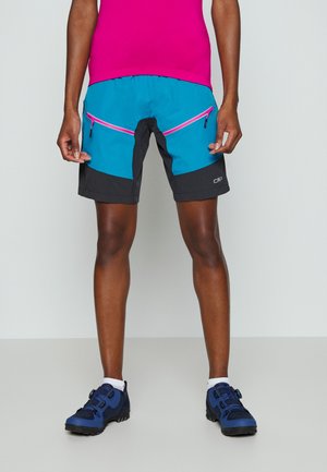 Personne portant un short de randonnée bleu et noir avec des poches zippées roses, un haut rose, des chaussettes blanches et des chaussures de randonnée bleues, debout contre un mur uni.