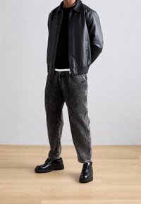 Zwarte leren bomberjack over een zwarte trui, gecombineerd met grijs gestreepte ribfluwelen broek en zwarte veterschoenen op een houten vloer.