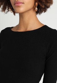 Haut noir côtelé avec un col rond, manches longues et tissu texturé. Des boucles d'oreilles créoles dorées complètent la tenue.