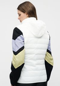 Γυναίκα με γυρισμένη πλάτη που φοράει ένα λευκό καπιτονέ μπουφάν με κουκούλα και μανίκια σε χρώματα μαύρο, ανοιχτό μωβ και μπεζ.