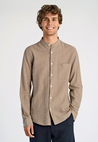 Camisa de color marrón claro, de manga larga, con cuello tipo banda, bolsillo frontal y botones blancos. La tela parece tener textura y es informal, combinada con pantalones oscuros.