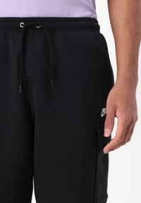 Calças de treino pretas feitas de algodão, com cintura elástica, cordão, bolso lateral e um pequeno logótipo branco da Nike na coxa.