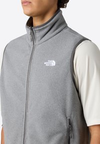 Person iført en lys grå fleecevest med lynlås og "The North Face"-logo på brystet over en hvid langærmet skjorte.