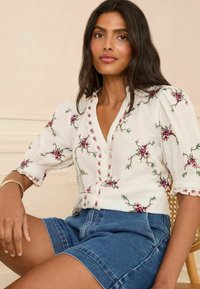 White blouse with floral embroidery featuring pink and green designs, short puff sleeves, and a buttoned front. Paired with denim shorts.