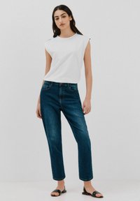 Hvid ærmeløs top parret med mørkeblå lige ben jeans, der har en glat stofstruktur og minimalistisk design. Sorte slide sandaler fuldender looket.