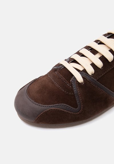 Καφέ σουέντ αθλητικό παπούτσι με σκούρα δερμάτινα στοιχεία και εκρού κορδόνια, εμφανιζόμενο από κοντινή γωνία σε λευκό φόντο.