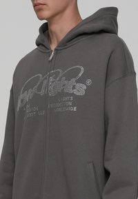 Sudadera con cremallera en gris, fabricada en un material suave, que presenta un logo frontal con adornos de cristales. Incluye capucha ajustable y bolsillos laterales.