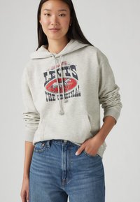 Grå hoodie med en framficka i kenguru-stil, med en tryckt logotyp i rött och svart. Kombinerad med högmidjade blå jeans som har en subtil textur.