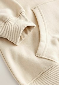 Cremefarbener Pullover mit Rippbündchen und -saum, flacher Taschenapplikation und glattem, strukturiertem Stoff.
