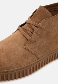 Stivale chukka in suede marrone con punta arrotondata, lace-up frontale e suola in gomma testurizzata con righe orizzontali.