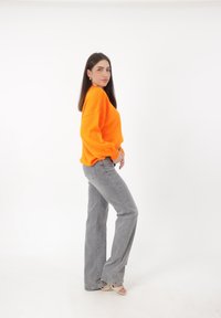 Heller orangefarbener Oversized-Pullover mit gerippten Säumen, kombiniert mit hellgrauen, ausgestellten Jeans und beigen Sandalen mit Riemchen. Einfarbiger weißer Hintergrund.