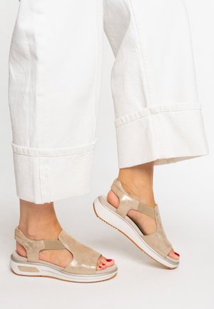 Beige suede sandals with open toes and elastic accents, featuring a white cushioned sole and textured detailing. Worn with white cropped pants.