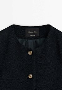 Svart texturerad kappa med rund halsringning, försedd med guldknappar och en slät innerfoder. Etikett med texten "Massimo Dutti, tillverkad i Rumänien."