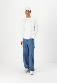 Chemise blanche à manches longues, jean cargo bleu et baskets blanches. La tenue est complétée par une casquette beige clair. Design simple aux tons neutres.