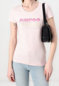 Kobieta w jasnoróżowej koszulce "GUESS Los Angeles", jasnoniebieskich dżinsach, z tatuażem na lewym ramieniu i czarną torbą na ramię na białym tle.