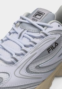 Λευκό και γκρι αθλητικό παπούτσι FILA με πλέγμα ύφασμα, ανακλαστικές ρίγες, παχιά μπεζ σόλα και υφαντά λευκά κορδόνια περασμένα από θηλιές.
