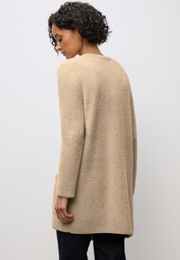 Cappotto beige realizzato in tessuto testurizzato, con maniche lunghe e tasche laterali. Presenta una vestibilità ampia e un orlo arrotondato. Visibile la vista posteriore.