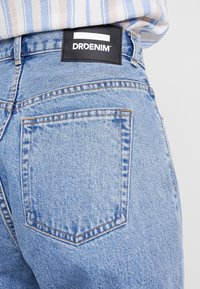 Blå denimjeans med en lätt tvätt. Har en rektangulär bakficka, synliga sömmar och en svart etikett med texten "DRDENIM."