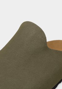 Κοντινή λήψη άνω μέρους παπουτσιού από σουέτ ελιάς με απαλή, καμπυλωτή σχεδίαση και ταμπά εσωτερική επένδυση ορατή στην άκρη.