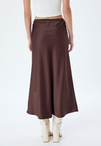Gonna in satin marrone con una silhouette a linea A fluida, caratterizzata da una texture liscia e una leggera lucentezza, indossata con stivali bianchi fino al ginocchio.