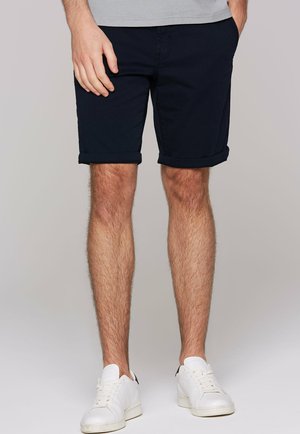Man wearing black rolled-up shorts, white sneakers, and a gray striped shirt with one hand in pocket.