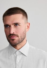 Light gray checked shirt with a button-down collar, textured fabric, and visible stitching details, worn by a person with short hair.