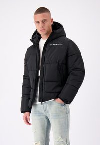 Zwarte pufferjack met een capuchon, voorzien van een rits aan de voorkant, zijzak en een wit logo op de borst. Het materiaal lijkt glad en geïsoleerd.