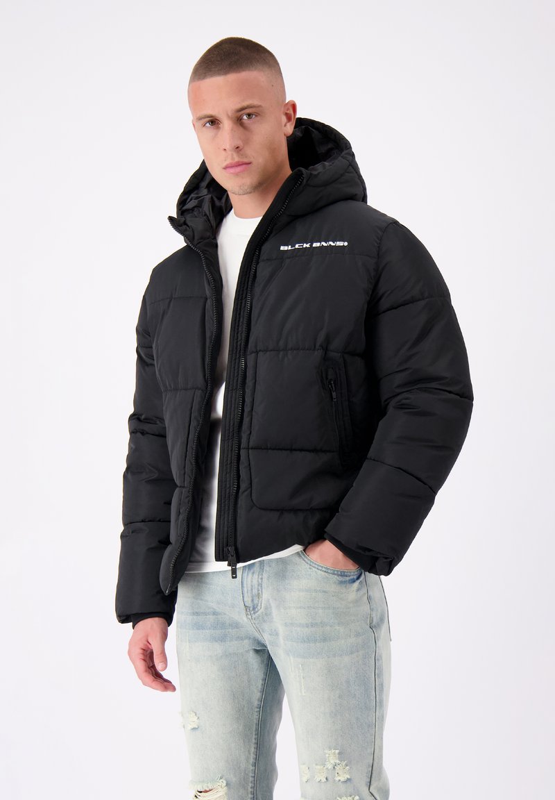 Zwarte pufferjack met een capuchon, voorzien van een rits aan de voorkant, zijzak en een wit logo op de borst. Het materiaal lijkt glad en geïsoleerd.