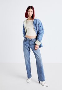 Jeansjack met schapenvacht kraag, beige crop top, blauwe high-waisted jeans en witte spitse hakken tegen een effen achtergrond.