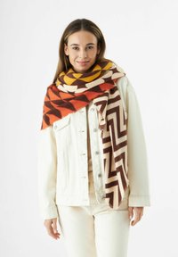 Jonge vrouw in een crème jack en broek met een grote kleurrijke geometrische sjaal om haar nek en schouders.