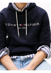 Námořnická mikina s kapucí a bílými stahovacími šňůrkami, s logem "TOMMY HILFIGER" a textem "EST. 1985". Nositá přes modro-bíle pruhovanou košili.