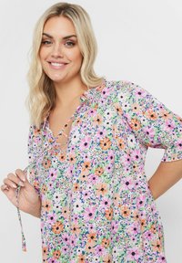 Blommig tunika med en mängd färgglada blommor på en ljus bakgrund. Viktiga designelement inkluderar en V-ringning och dragsnöre.