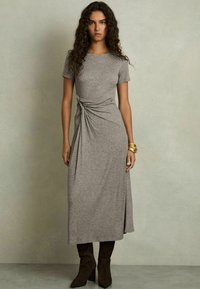 Šedý jersey šaty s okrúhlym výstrihom, krátkymi rukávmi a bočným twist detailom; majú dlhý priliehavý siluet. Hnedé podpätkové čižmy.