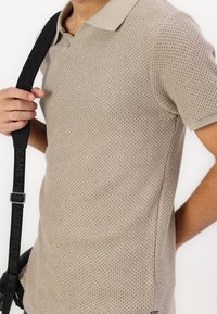 Polo beige avec un motif texturé en nid d'abeille et un col contrastant. Le modèle tient un sac à bandoulière noir avec des détails de logo.
