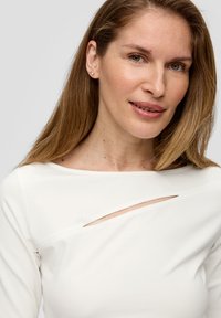 Witte lange mouwen top met een diagonale uitsnijding over de borst, een gladde textuur en een aansluitend ontwerp. Het haar is stijl en van gemiddelde lengte.